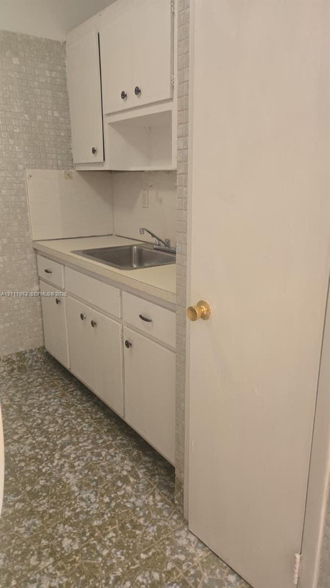 Kitchenette featuring white cabinetry, a neutral-toned countertop, a stainless steel sink with a chrome faucet, mosaic wall tiling, and patterned tile flooring