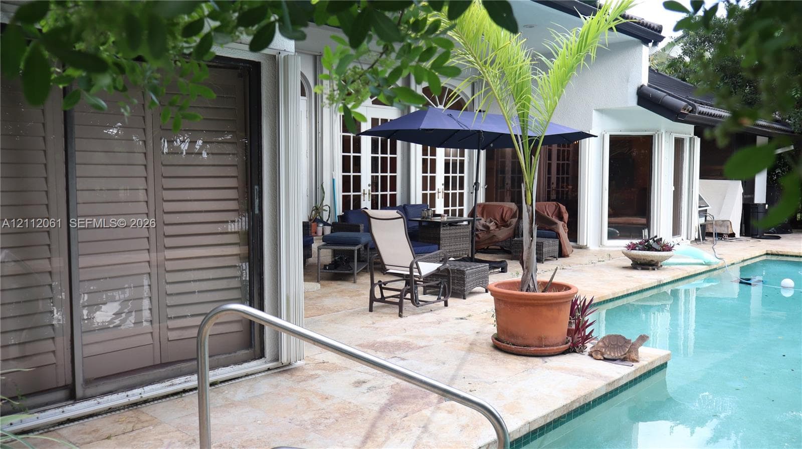Swimming pool with a metal handrail, light-toned stone patio, multiple sets of French doors, and exterior sliding shutters