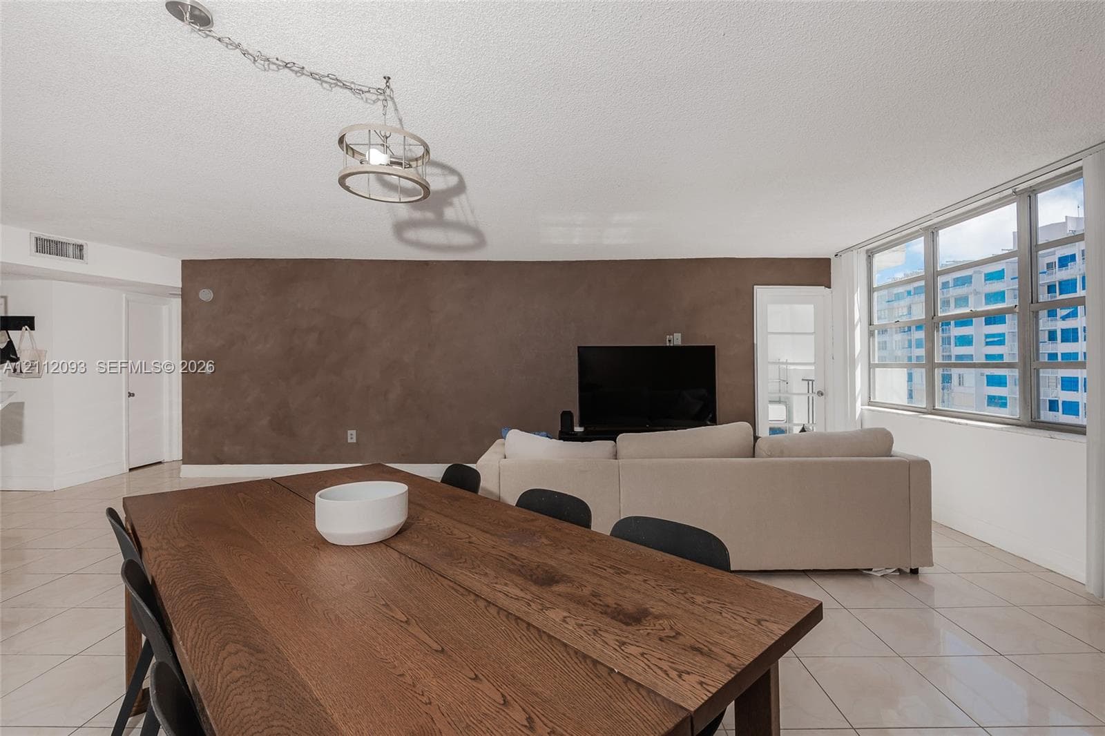 Open-concept living space featuring large-format ceramic tile flooring, a prominent accent wall with textured finish, expansive multi-pane windows, and a suspended industrial-style drum pendant light fixture