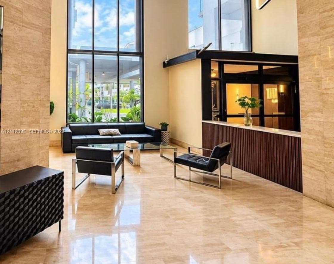 Double-height ceilings with floor-to-ceiling windows, polished stone flooring, stone-clad accent walls, and a built-in reception desk with vertical wood-slat detailing