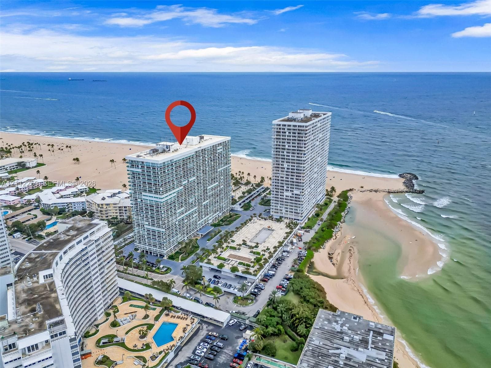 High-rise residential building featuring direct oceanfront positioning, ground-level swimming pool, paved surface parking, and manicured landscaping
