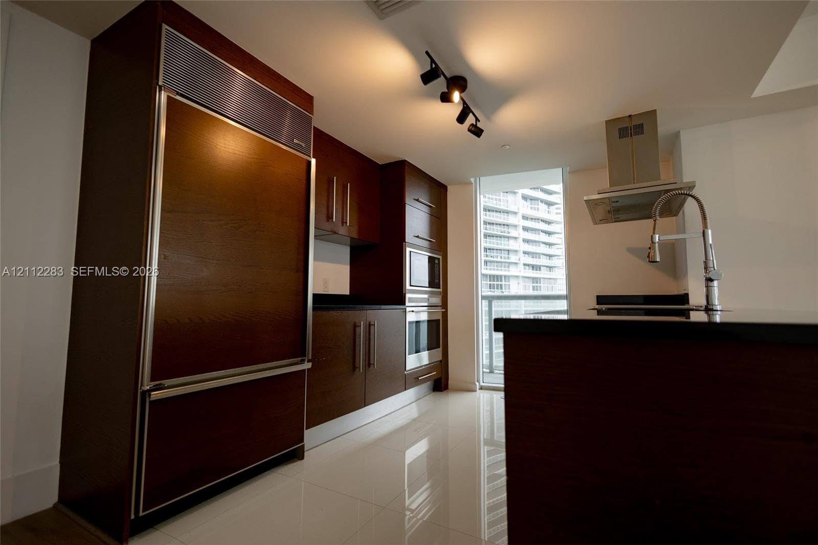 Kitchen featuring dark wood-finish cabinetry, stainless steel built-in appliances, a center island with a stone countertop, a gooseneck faucet, and high-gloss tile flooring