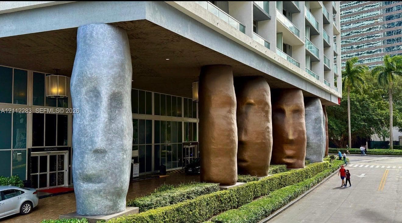 Exterior facade featuring large-scale sculptural columns, floor-to-ceiling glass windows, recessed entry lighting, and structured perimeter hedging