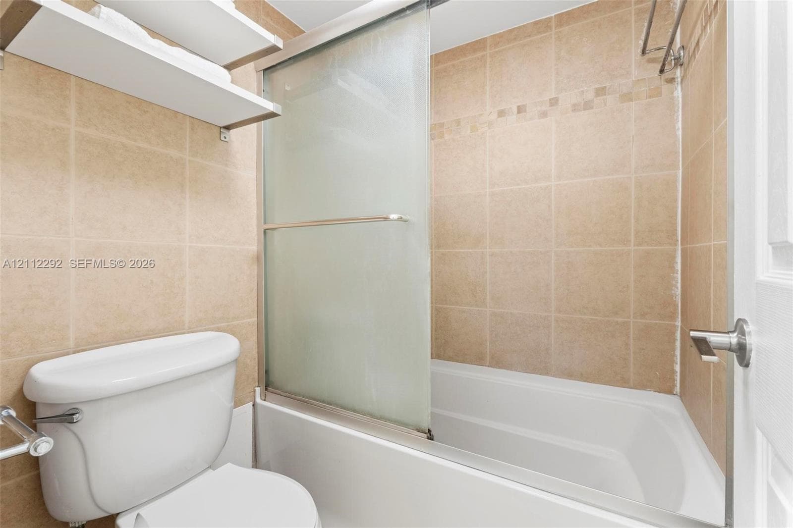 Bathtub enclosure with a sliding frosted glass door, tile surround, wall-mounted shelving, and polished metal hardware