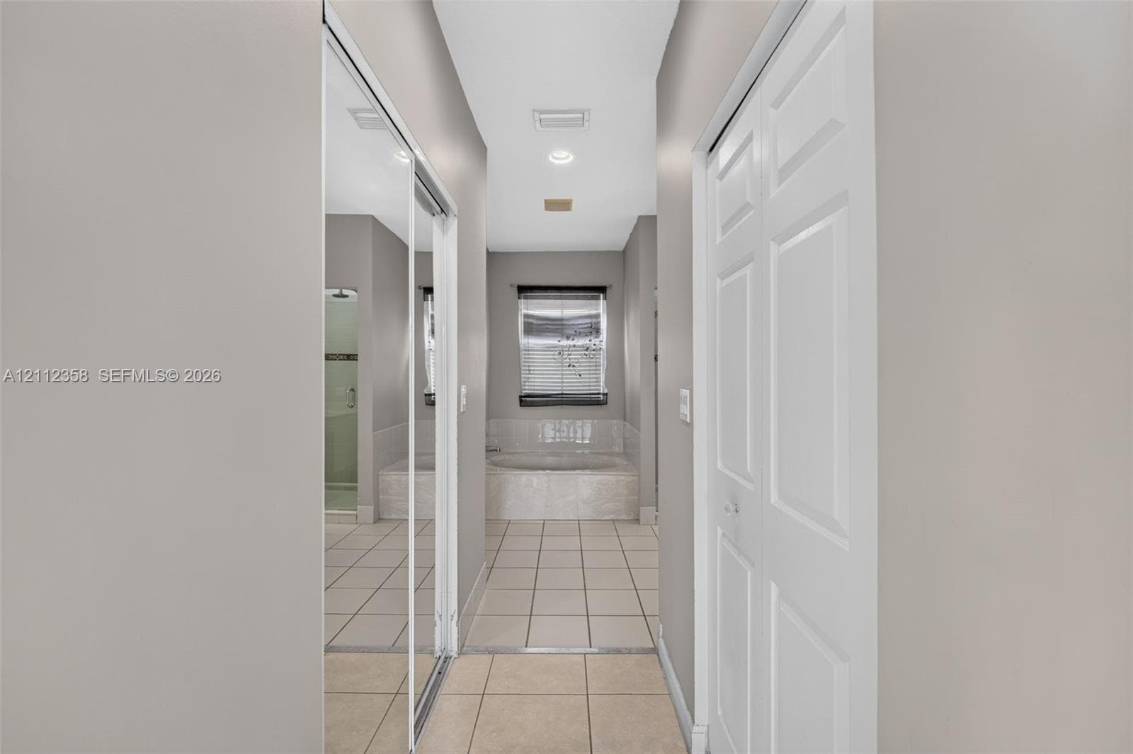 Hallway featuring mirrored sliding closet doors, white six-panel door, recessed lighting, and tile flooring with access to a tile-surround soaking tub