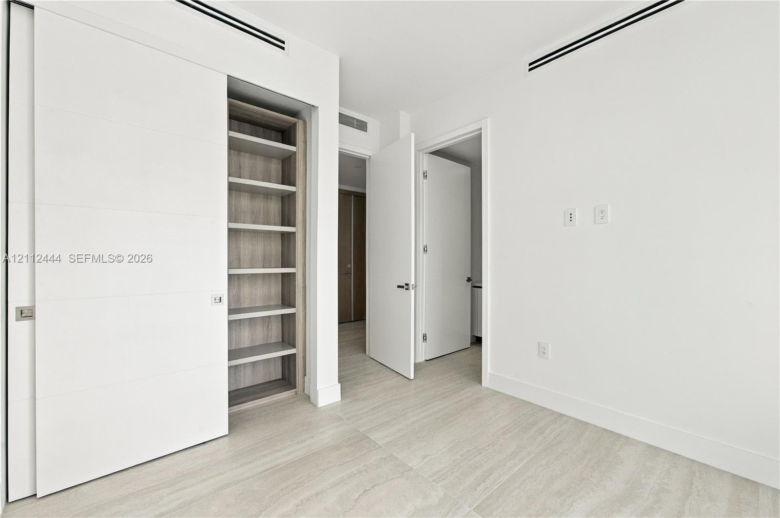 Minimalist interior featuring wood-finish flooring, recessed linear ceiling diffusers, sliding panel doors, and built-in shelving with wood-tone interior