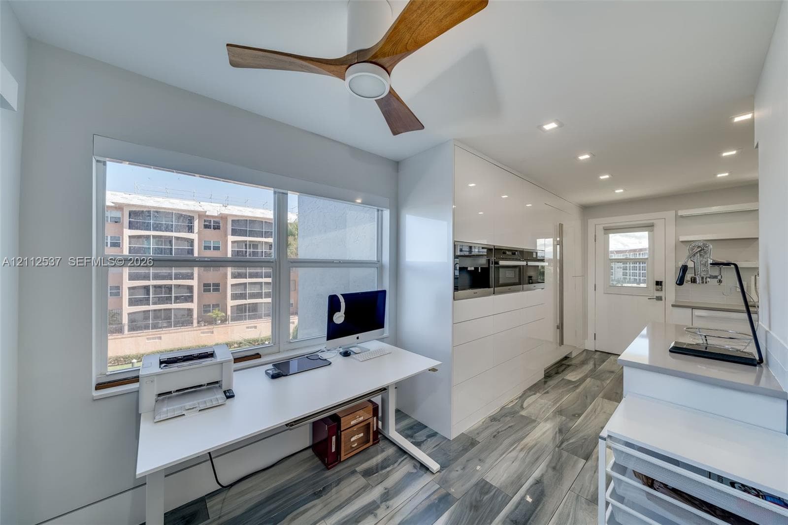Modern kitchen featuring high-gloss white cabinetry, integrated stainless steel appliances, wood-finish ceiling fan, recessed lighting, and large-format gray patterned flooring