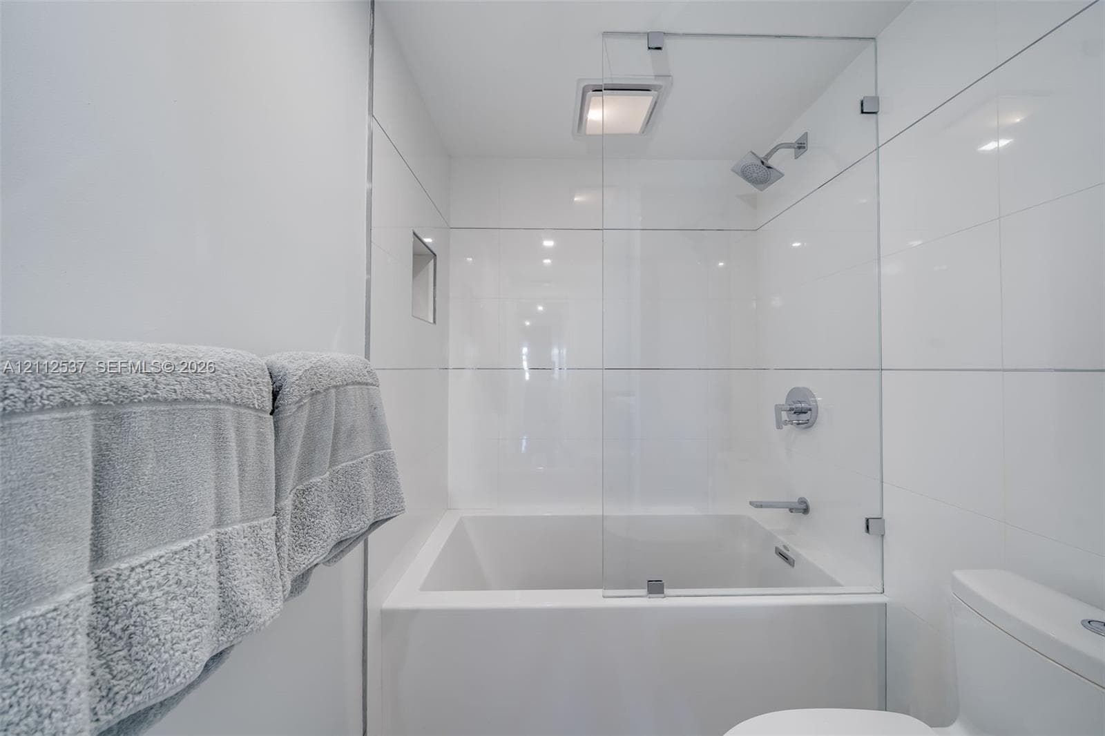 Soaking tub with a frameless glass splash panel, chrome rainfall showerhead, recessed wall niche, and floor-to-ceiling white tile surround