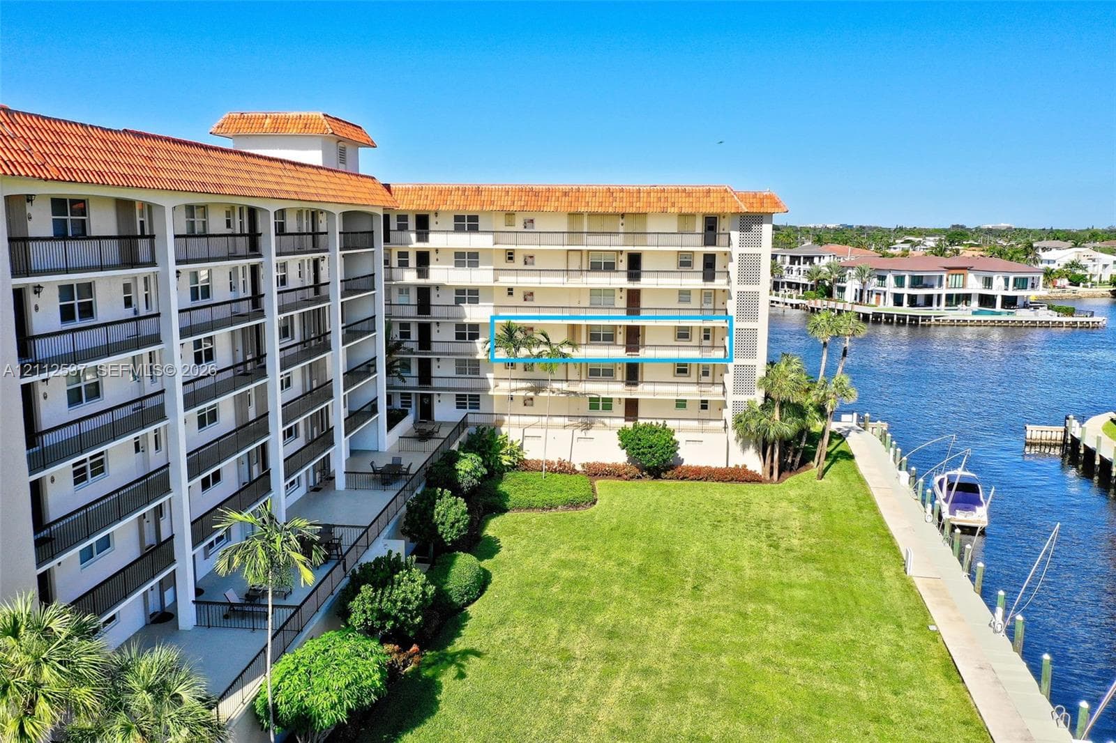 Multi-story residential building featuring a terracotta tile roof, private balconies with metal railings, and waterfront grounds with a manicured lawn