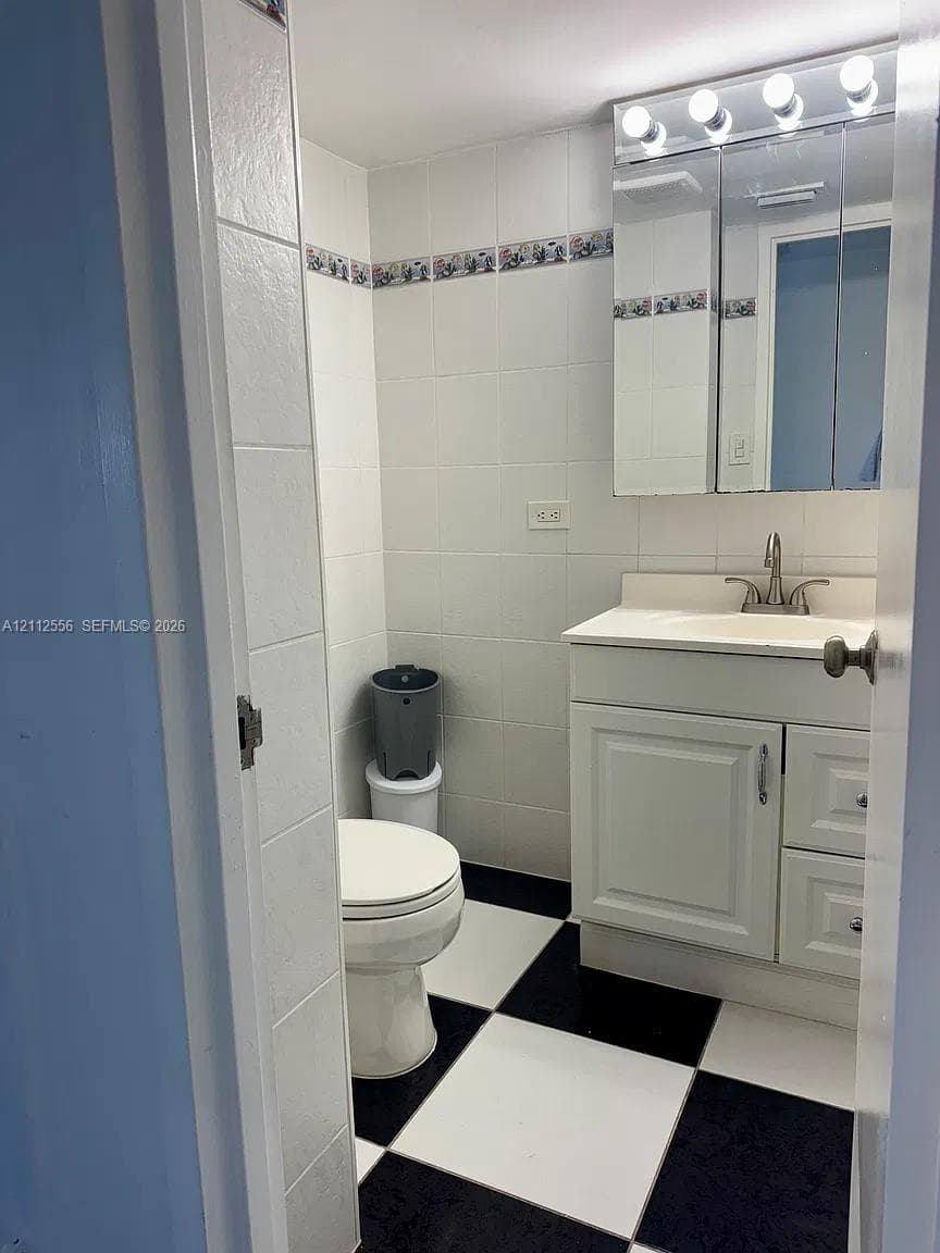 Bathroom featuring black-and-white checkered tile flooring, white wall tiling with decorative border, vanity cabinet with integrated sink, and mirrored medicine cabinet with overhead lighting
