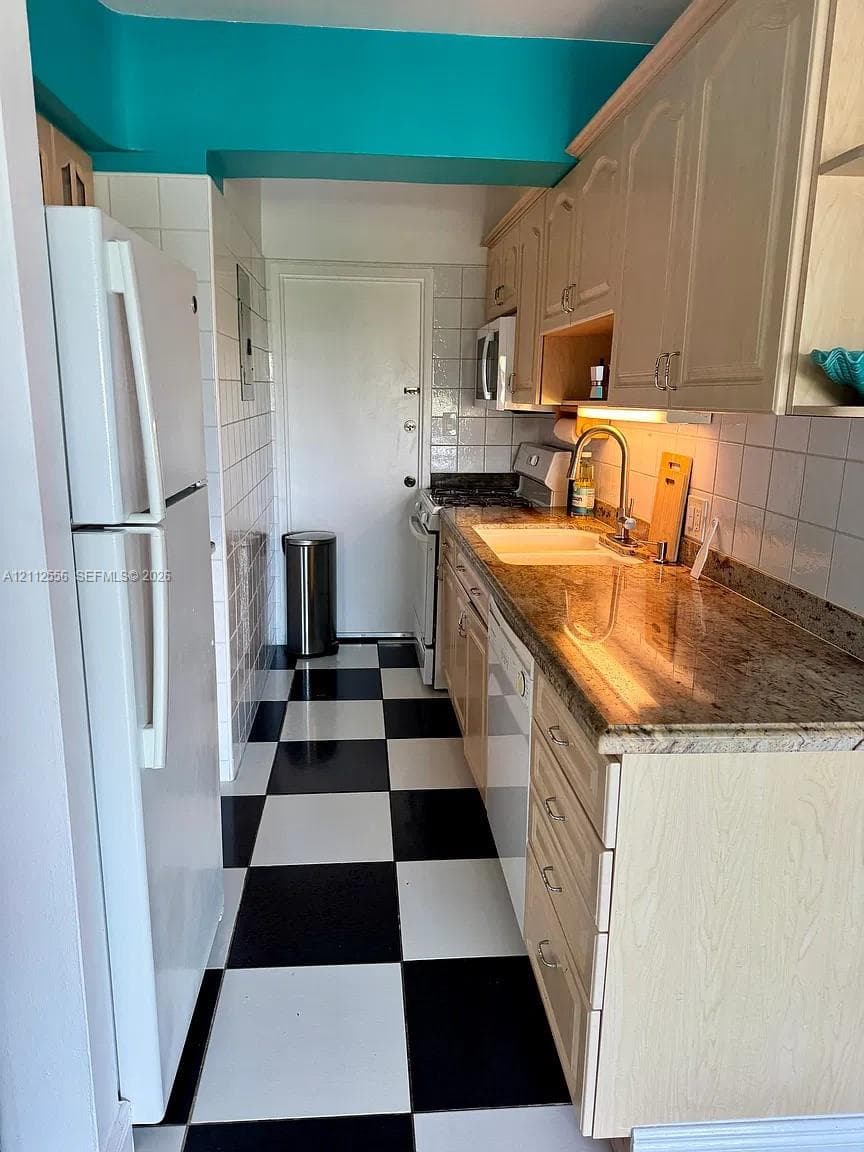 Galley kitchen featuring stone countertops, white tile backsplash, light wood-tone cabinetry, and checkered tile flooring