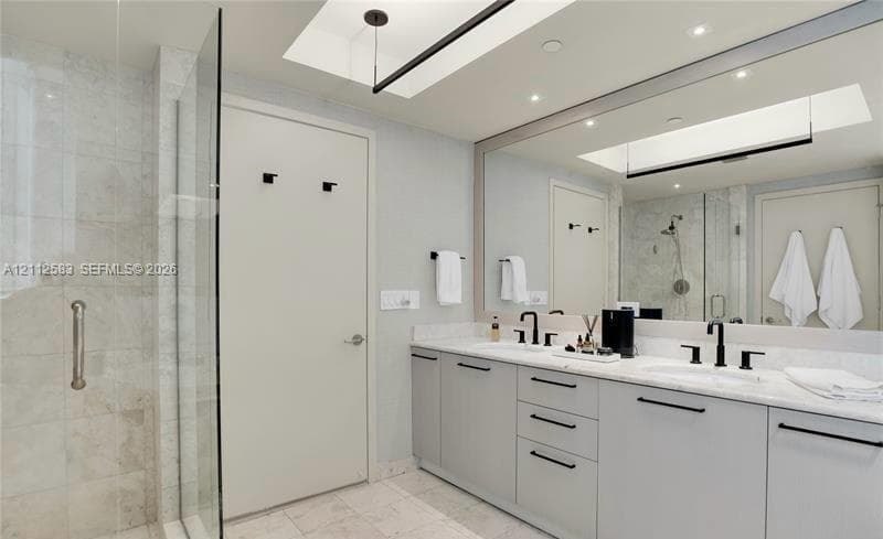 Dual-sink vanity with light-gray cabinetry, stone countertops, matte black fixtures, full-width mirror, and walk-in tiled shower with a glass enclosure