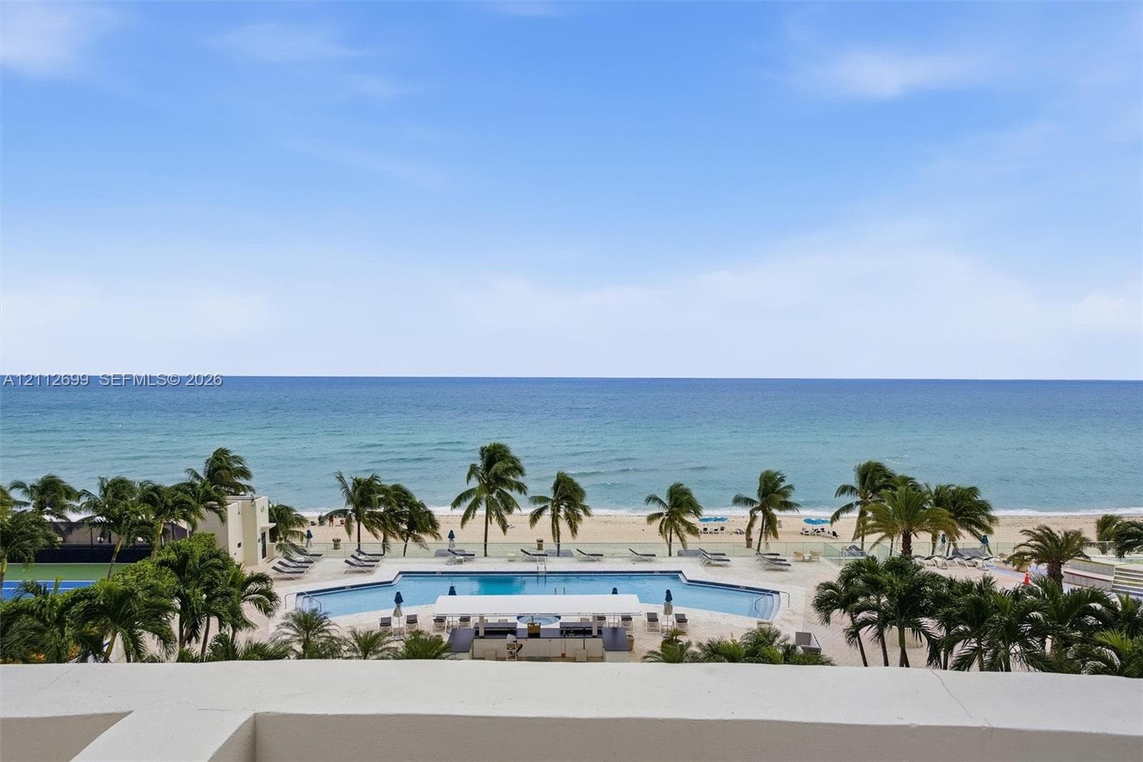 Oceanfront terrace featuring a concrete parapet, overlooking a resort-style swimming pool, lush palm trees, and sand beach
