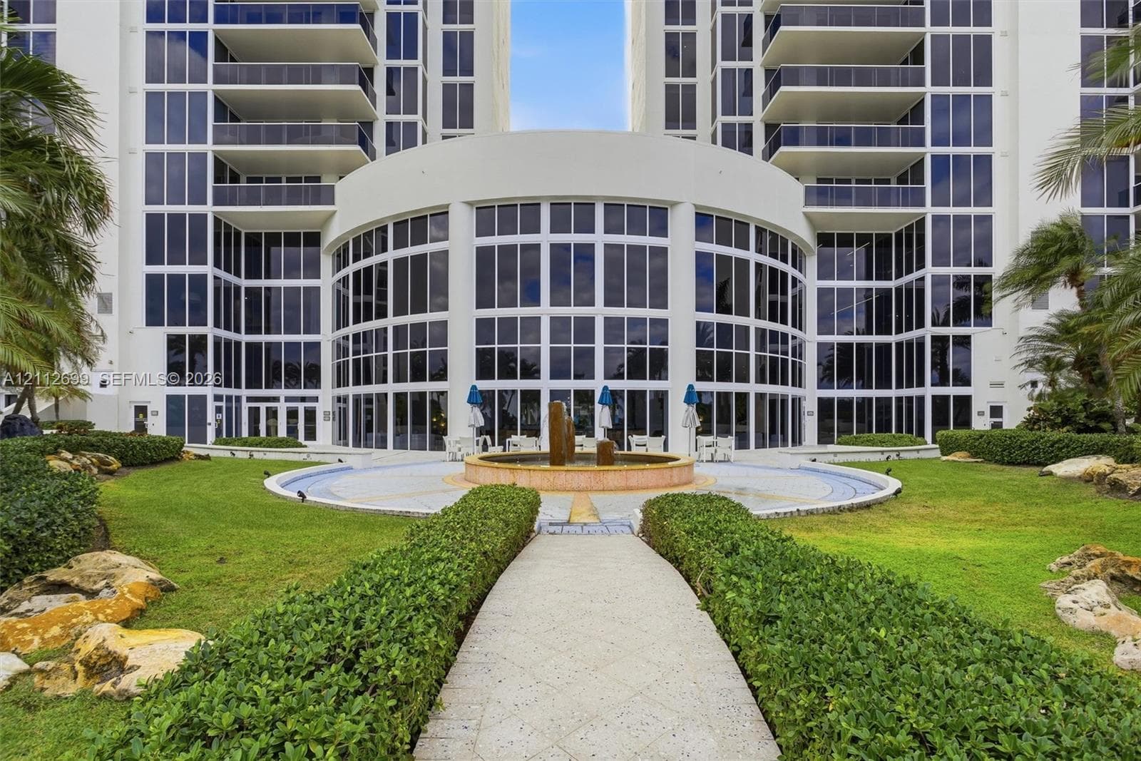 High-rise exterior featuring a multi-story glass facade, private balconies, a central water fountain, stone-paved pathways, and manicured landscaping