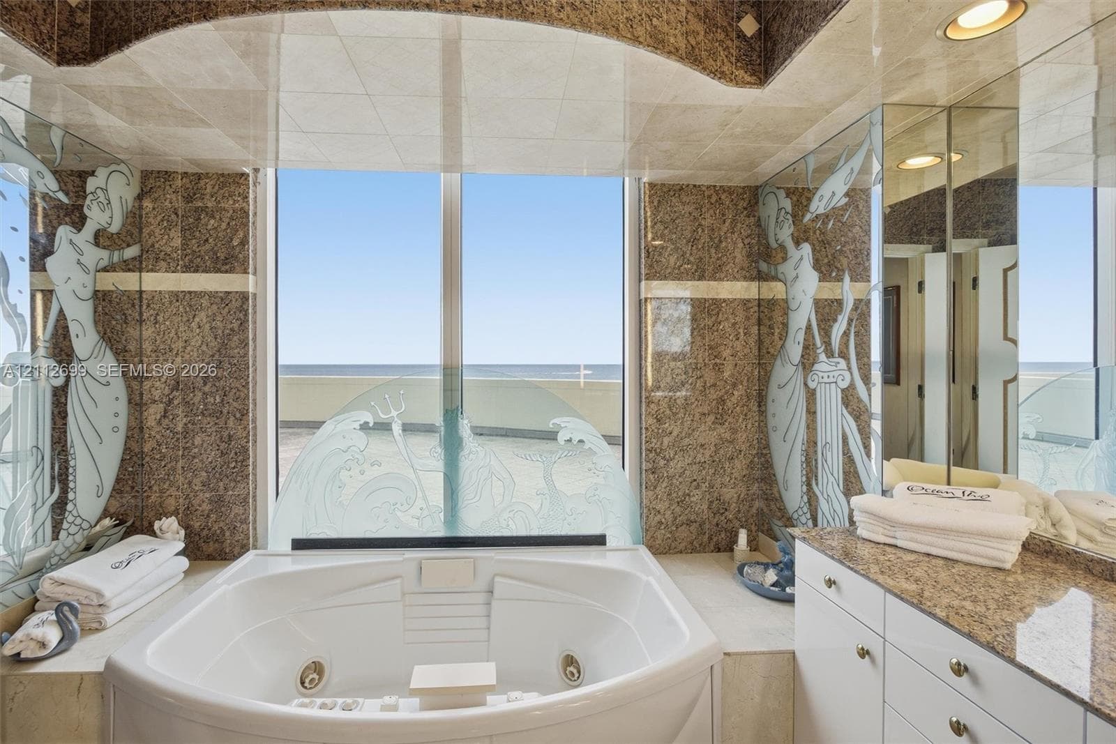 Custom soaking tub with integrated jets, large window with ocean horizon backdrop, stone wall cladding, decorative etched glass panels, and built-in vanity with stone countertop