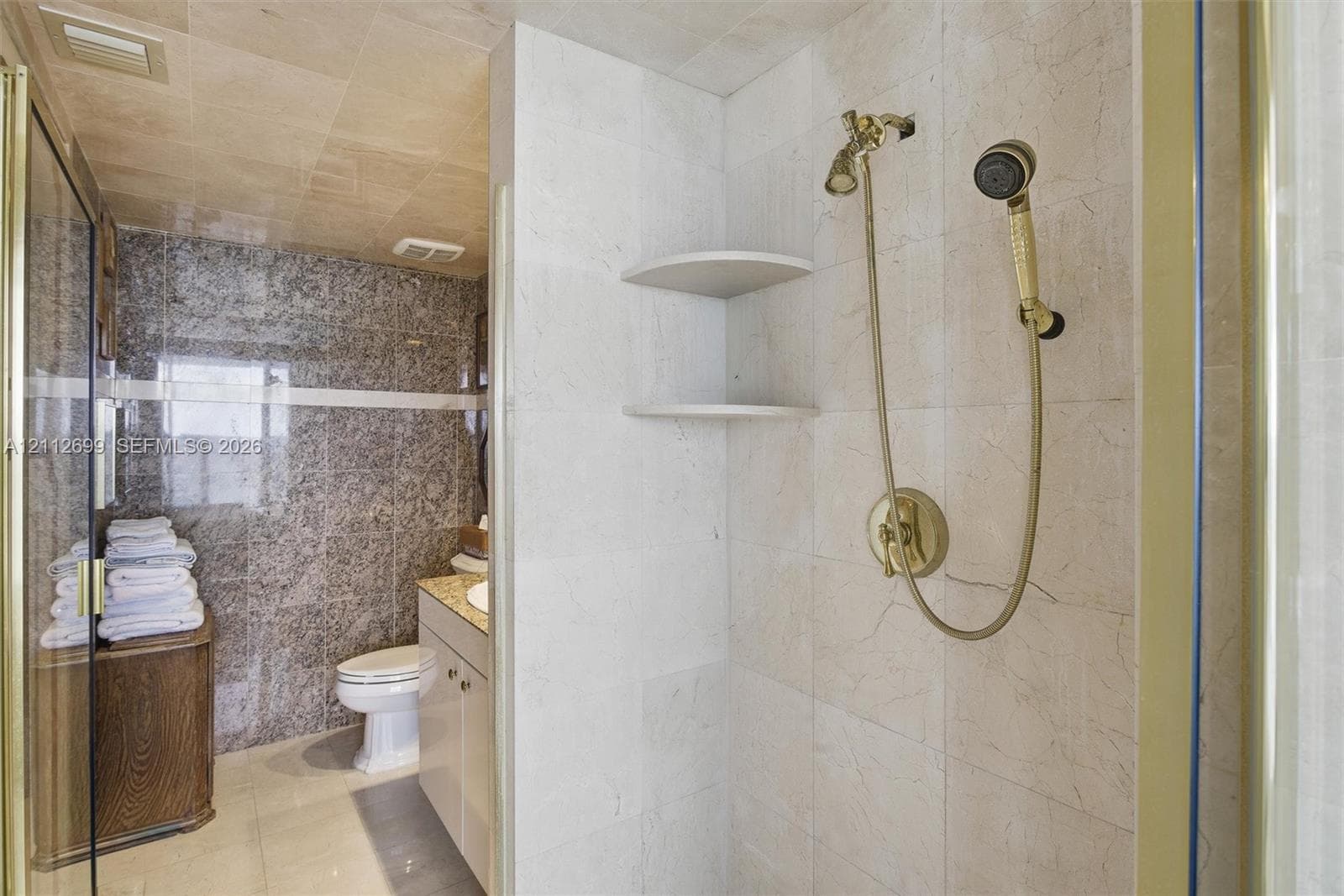 Walk-in shower enclosure featuring floor-to-ceiling neutral tile, built-in shelving, and gold-finish hardware including a handheld showerhead