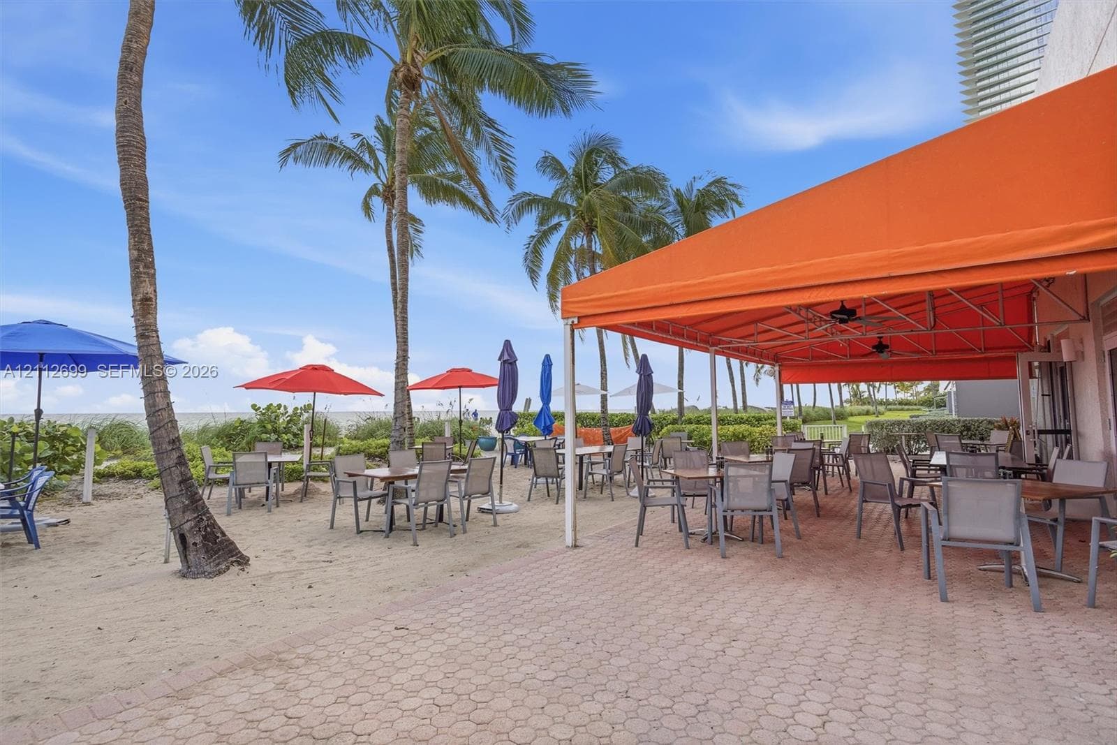 Expansive paver patio featuring a large fixed orange awning, direct sandy beach access, mature palm trees, and ceiling-mounted fans
