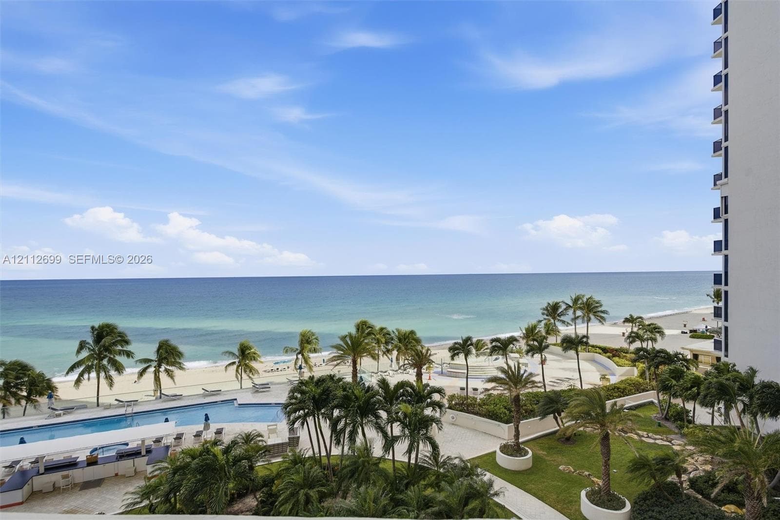 Elevated perspective featuring direct oceanfront access, a sprawling swimming pool with surrounding terrace, landscaped grounds with mature palms, and expansive sand shoreline