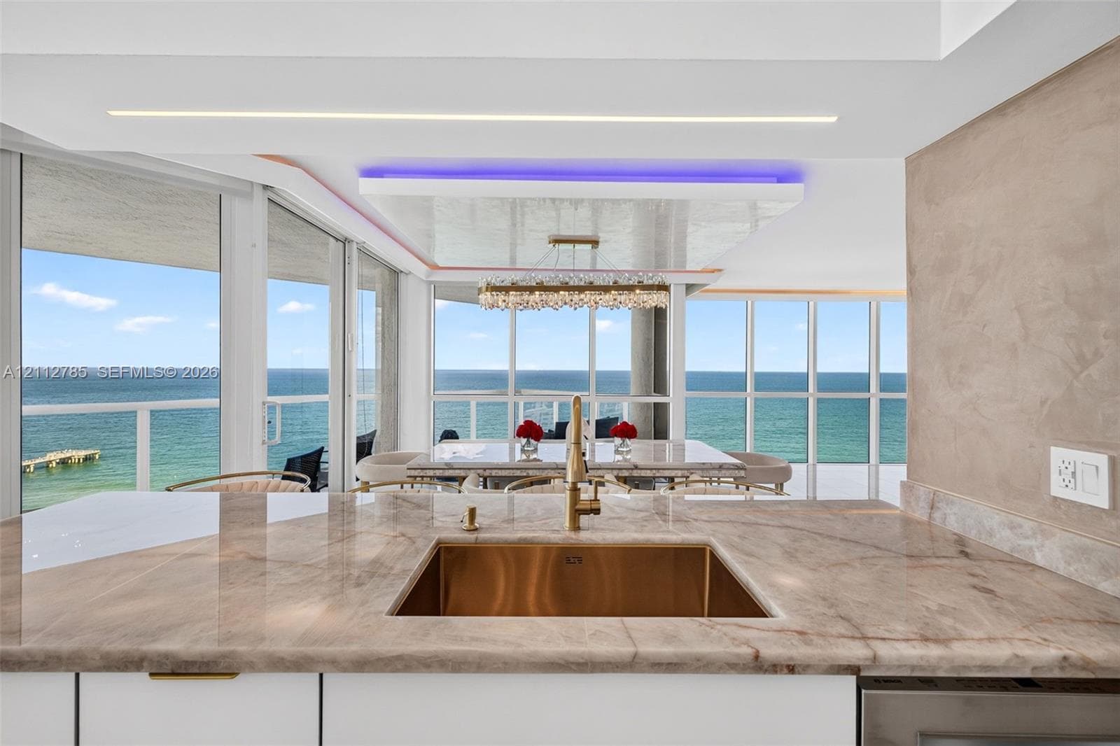 Kitchen featuring a polished stone countertop, undermount metallic basin, brushed gold-tone faucet, expansive floor-to-ceiling glass windows, and a coffered ceiling with integrated accent lighting
