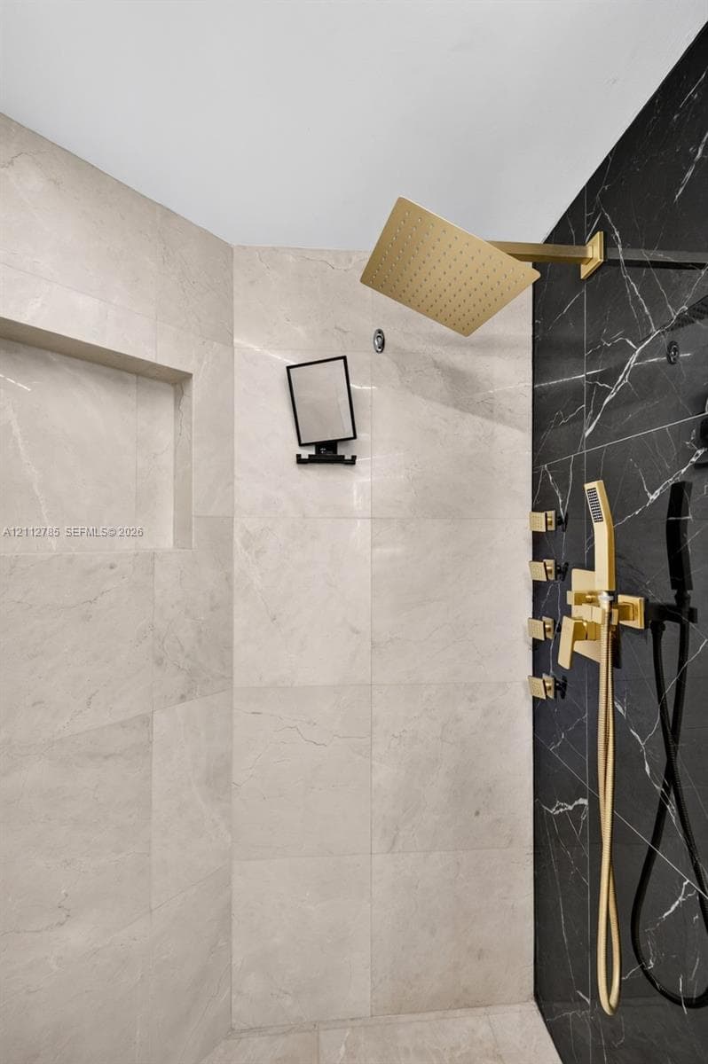 Walk-in shower featuring large-format light-toned tile, contrasting black tile accent wall, gold-finish rainfall showerhead, multi-body spray system, and integrated recessed shelving