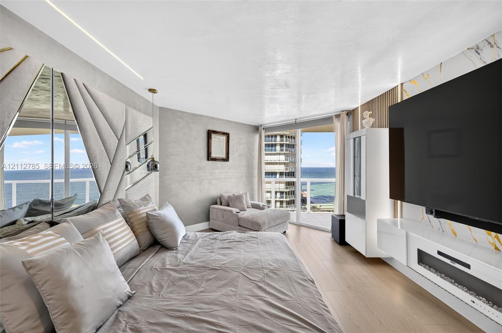 Wood-finish flooring, floor-to-ceiling windows with coastal scenery, accent wall with decorative mirror panels, wall-mounted linear fireplace, and integrated wood-slat wall paneling