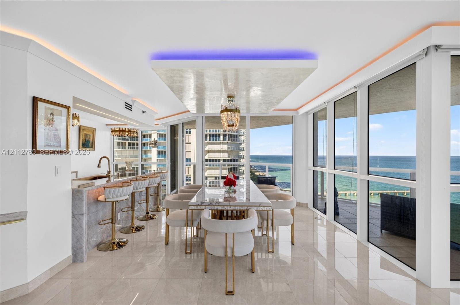 Floor-to-ceiling windows with ocean exposure, polished stone flooring, custom coffered ceiling with integrated accent lighting, wet bar with stone-surround, and a crystal chandelier