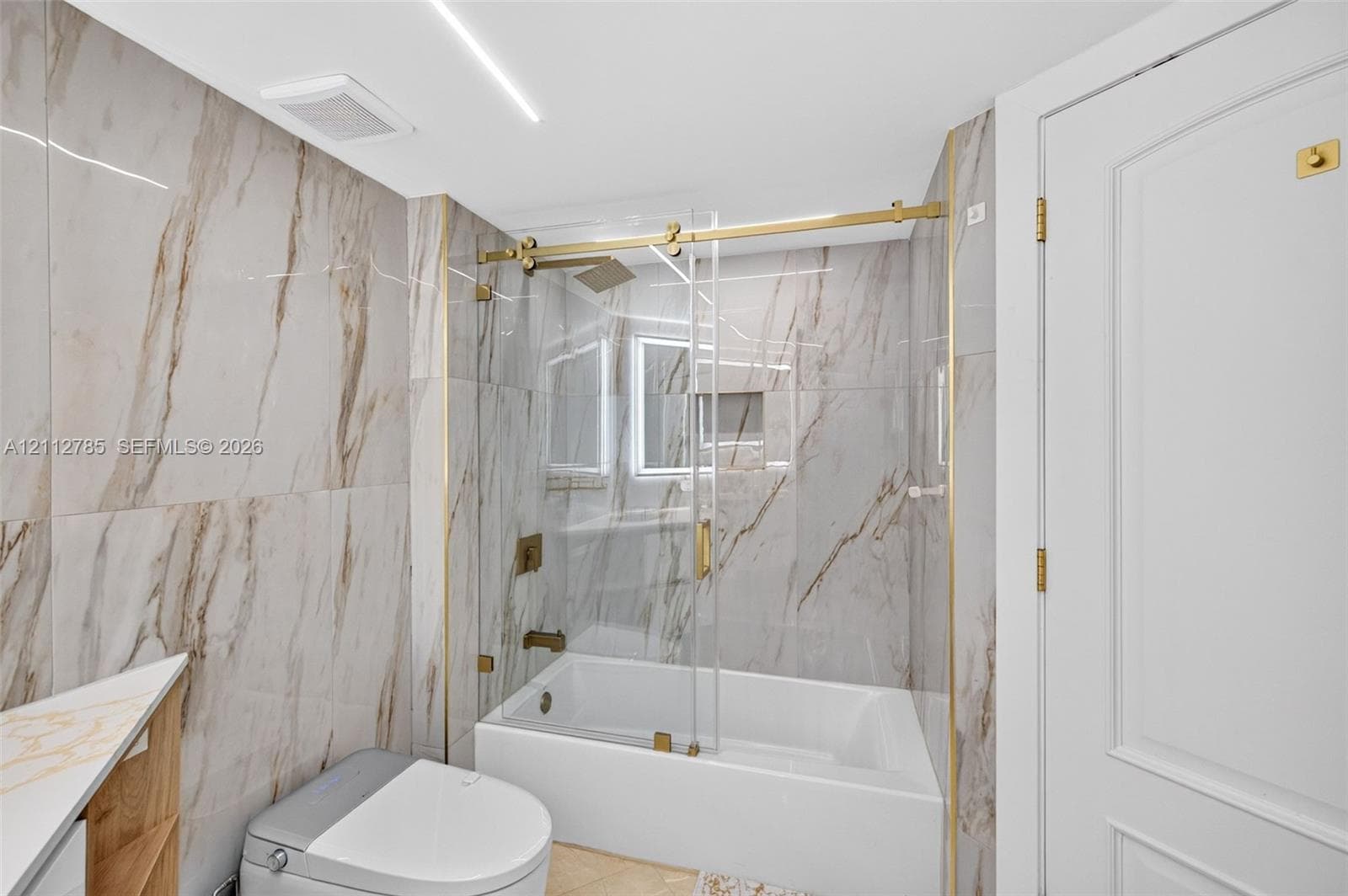 Full-height wall tiling with a vein-patterned finish, glass-enclosed shower with gold-toned hardware, built-in recessed wall niche, and modern white cabinetry