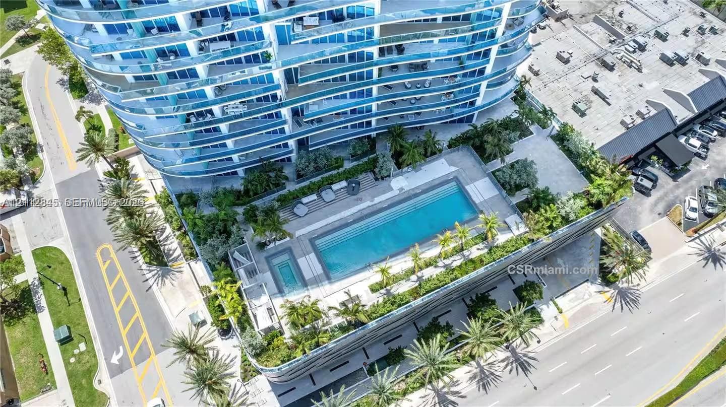 High-rise building featuring curvilinear architecture with glass balcony railings, an expansive ground-level swimming pool, a dedicated spa, and integrated tropical landscaping