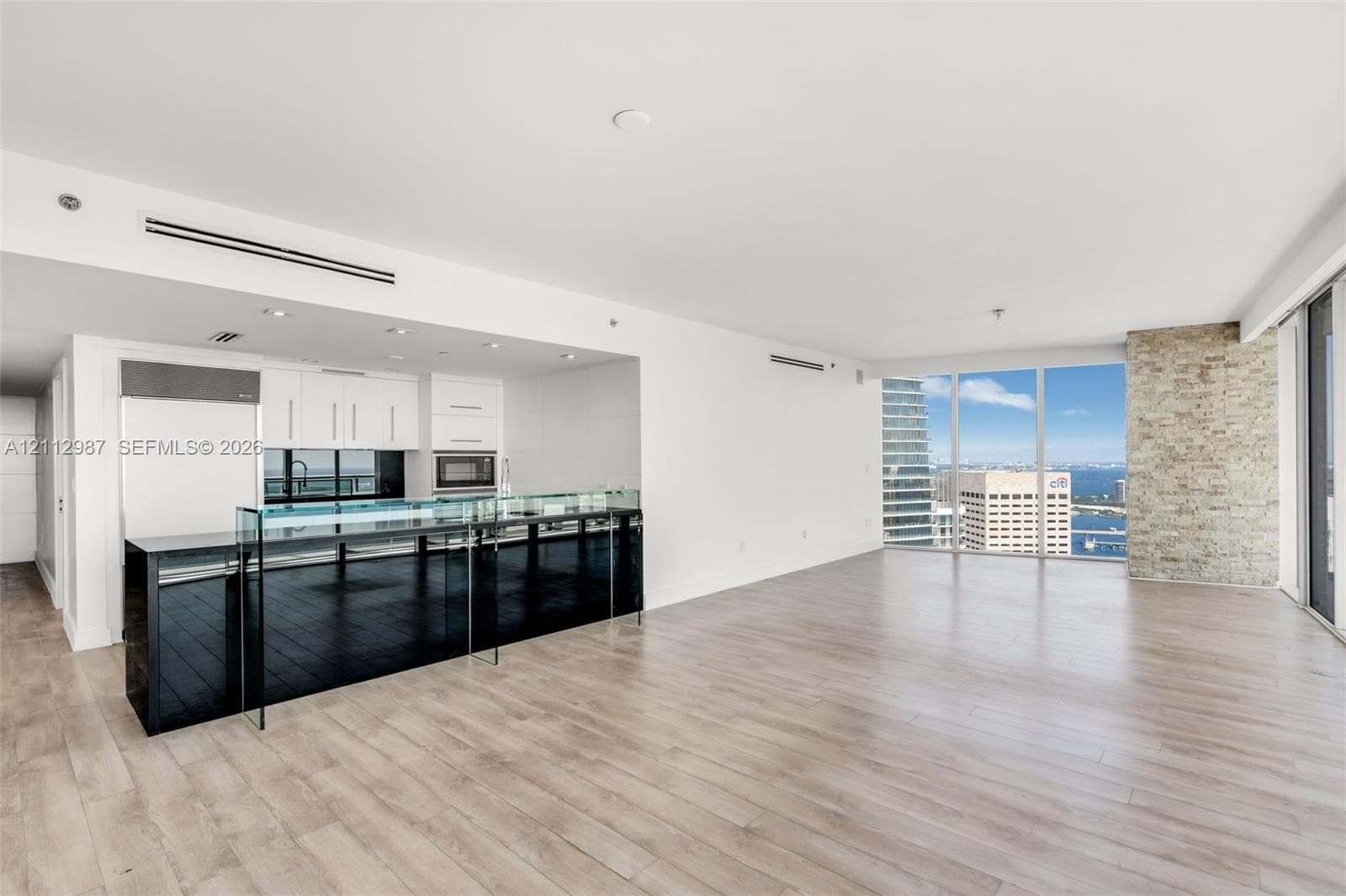 Open-concept kitchen featuring a black stone island with a glass breakfast bar, white cabinetry, integrated stainless steel appliances, wood-finish flooring, and a floor-to-ceiling brick accent wall
