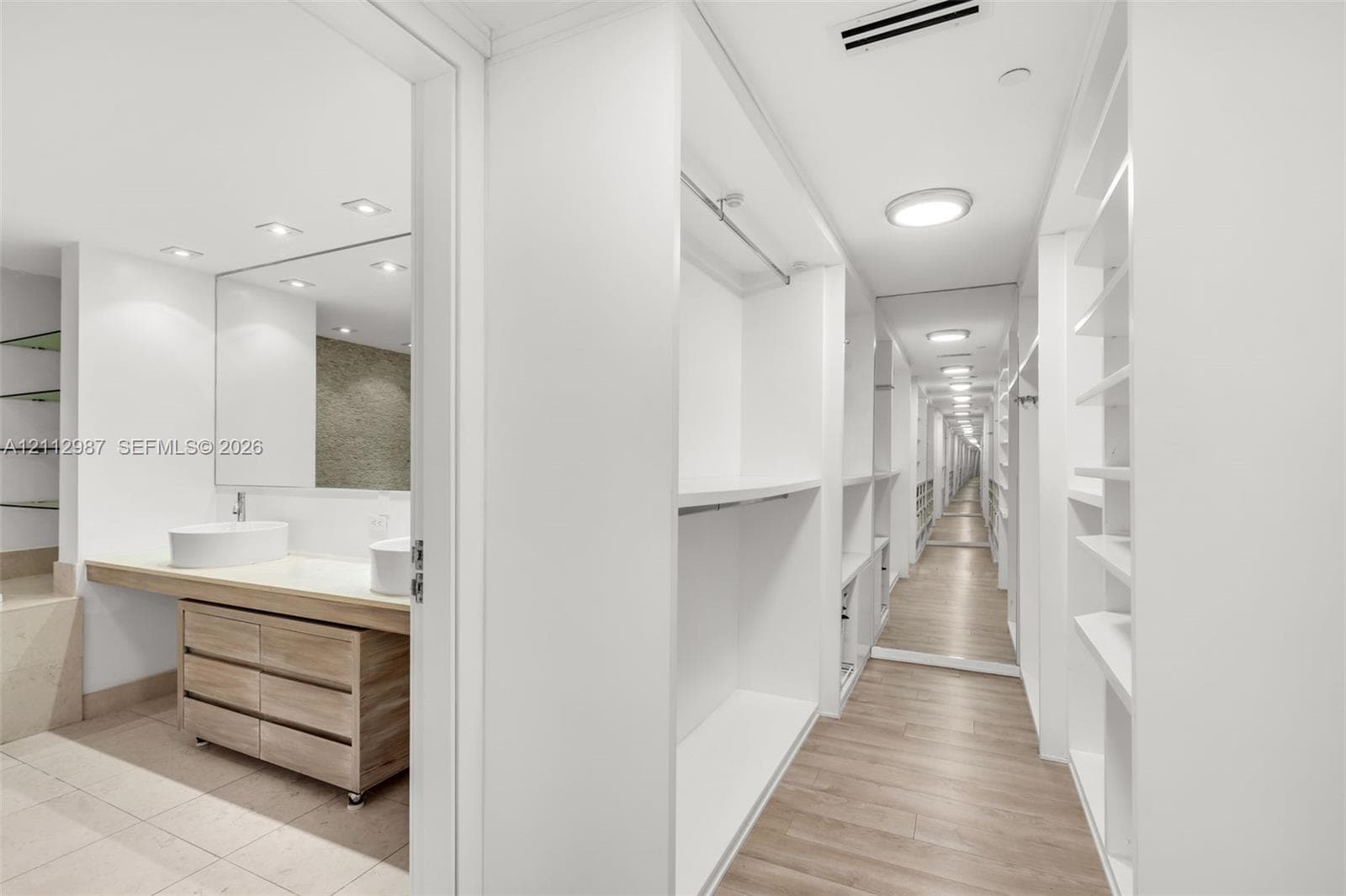 Walk-in closet featuring white built-in shelving, full-length mirror, and wood-finish flooring, accompanied by a bathroom with a dual-vessel sink vanity and neutral tile flooring