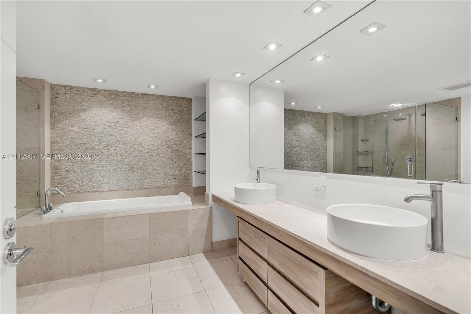 Bathroom featuring dual vessel sinks, a custom vanity with wood-finish cabinetry, a textured stone accent wall, a soaking tub, and a frameless glass shower enclosure