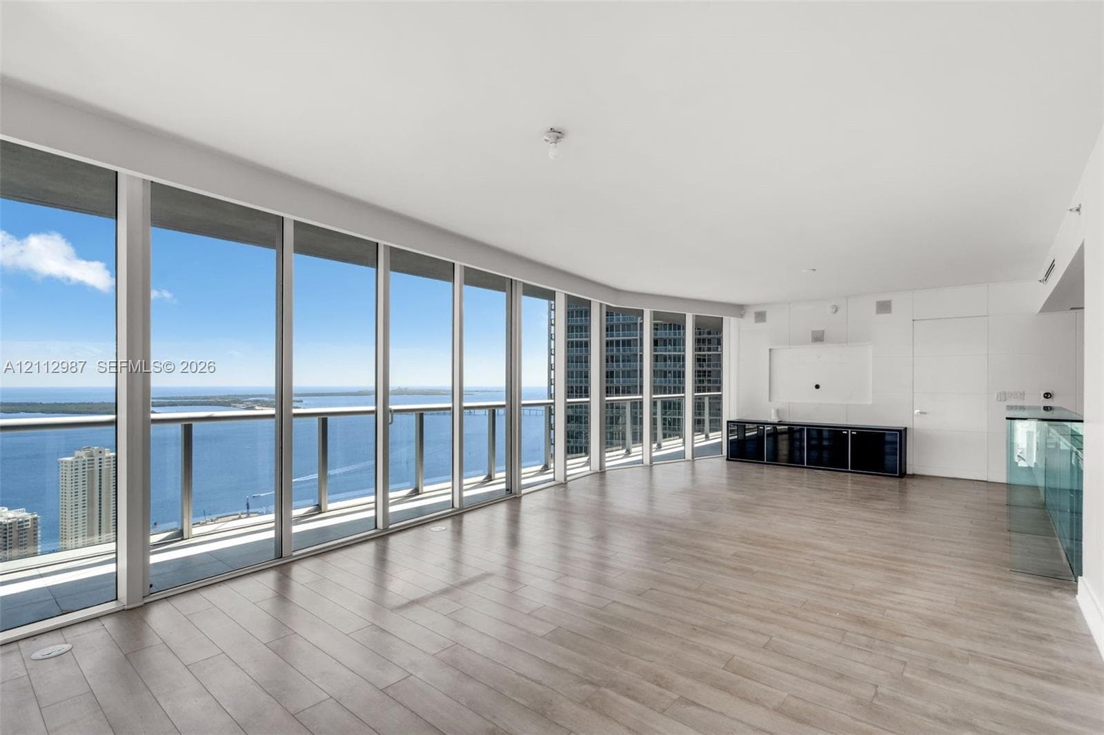 Floor-to-ceiling glass windows, wide-plank wood-finish flooring, and glass-railing balcony featuring expansive water and city skyline panoramas
