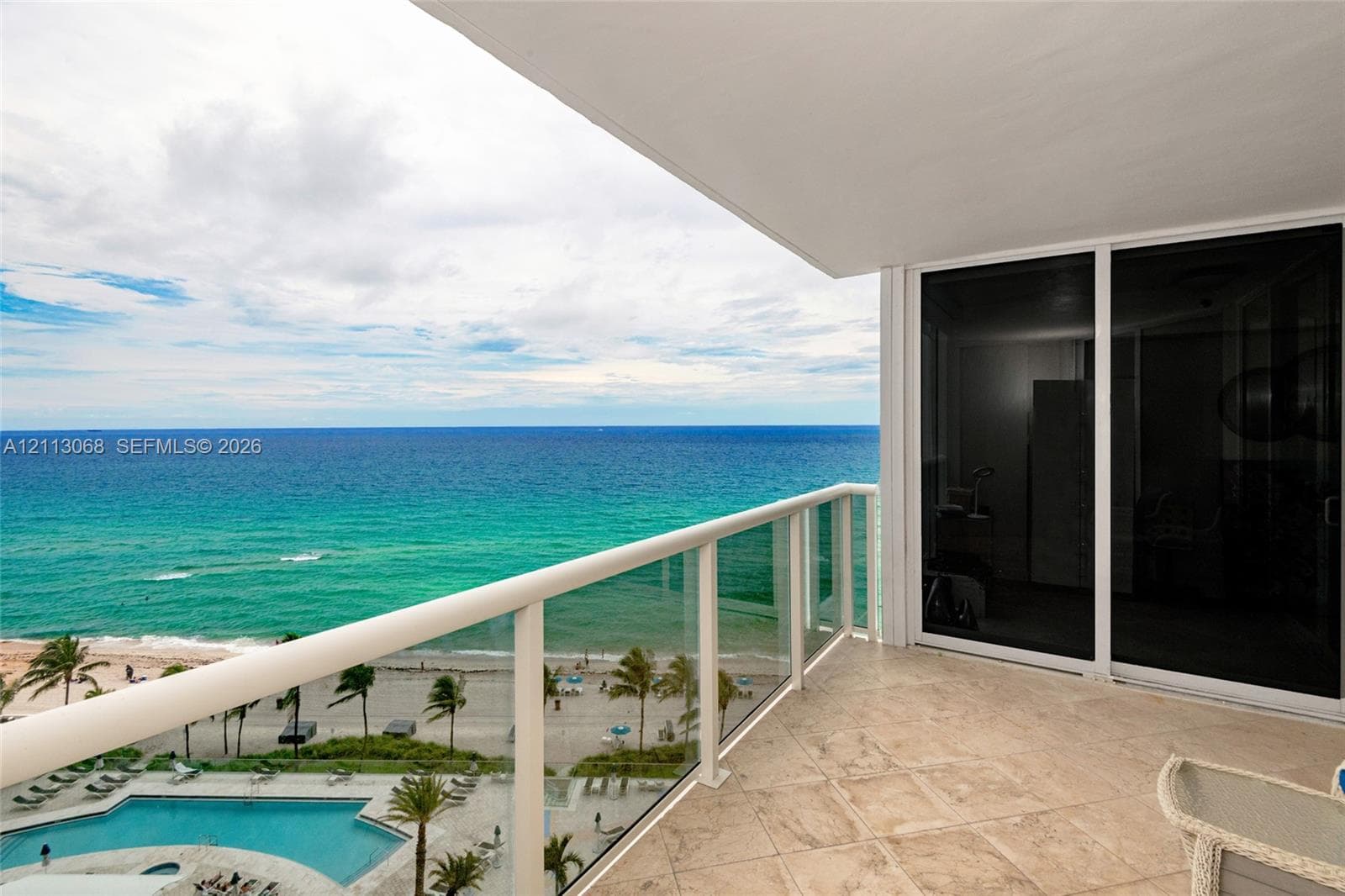 Covered balcony featuring stone flooring and a glass-paneled railing, with sliding glass doors and an oceanfront horizon
