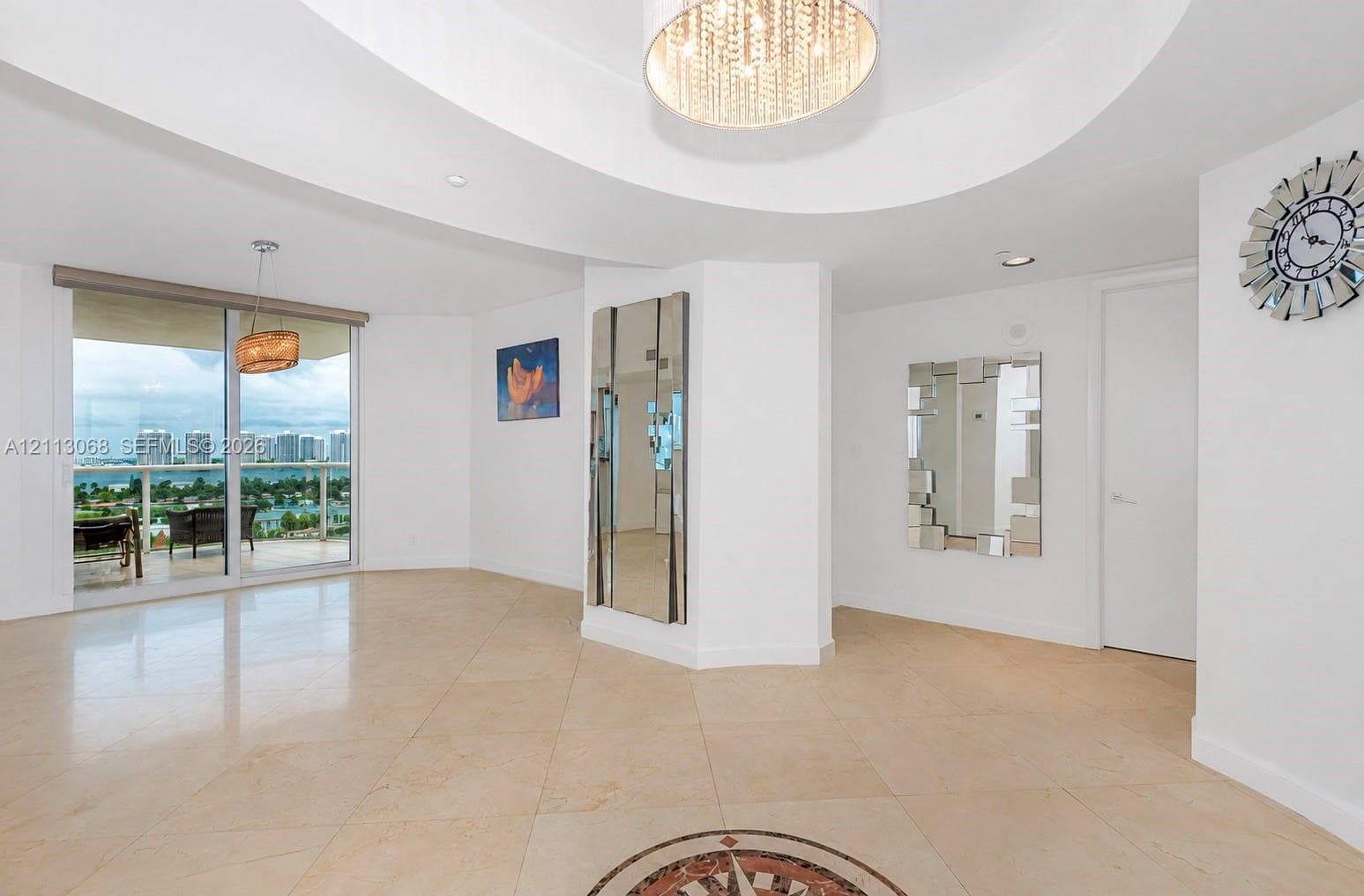 Open-concept interior featuring large-format tile flooring with a central medallion inlay, recessed ceiling architectural detailing, a tiered crystal drum chandelier, and a floor-to-ceiling mirror-paneled pillar