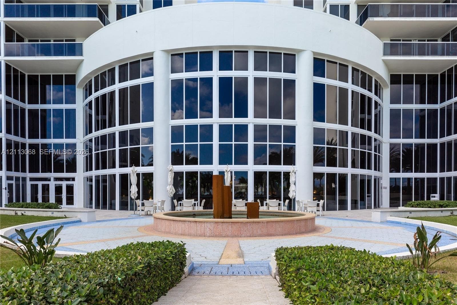 High-rise exterior featuring a multi-story glass curtain wall, rounded architectural facade, tiered stone fountain, and paved circular plaza