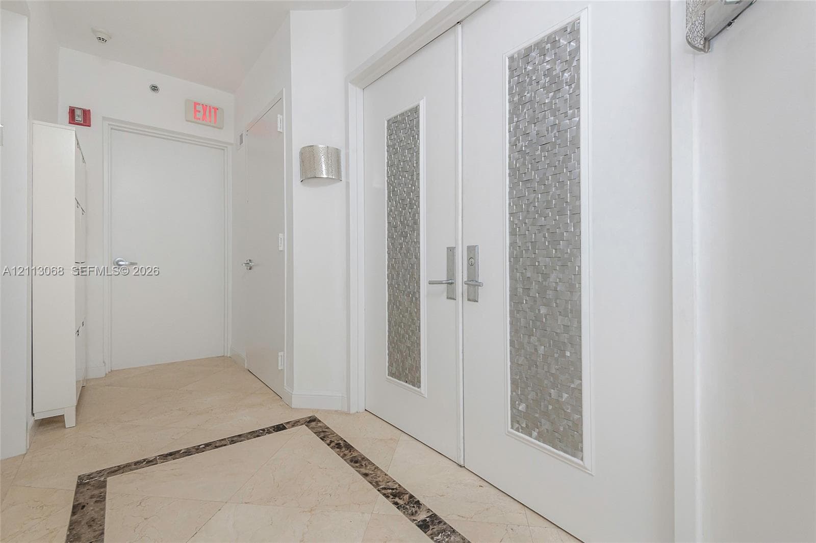 Double-entry doors featuring vertical textured metallic inlays, stone tile flooring with decorative stone border inlay, and wall-mounted sconce lighting