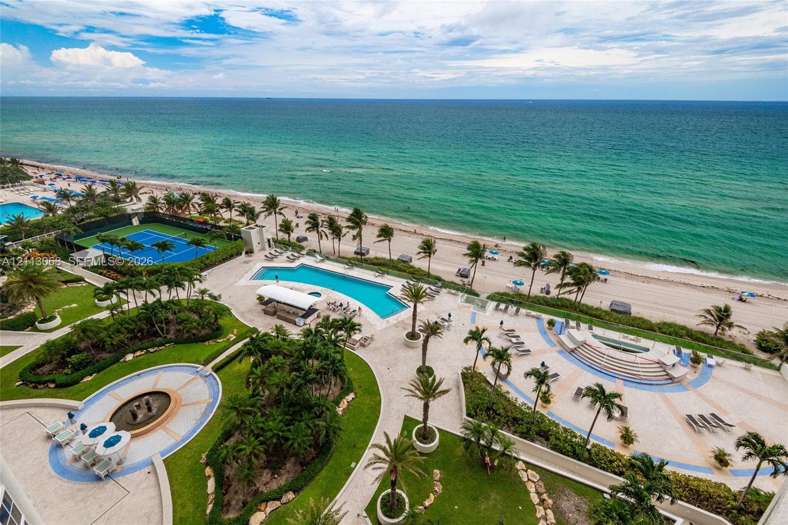Oceanfront complex featuring a swimming pool, tennis court, tiered stone patio, and circular fountain set amidst landscaped grounds
