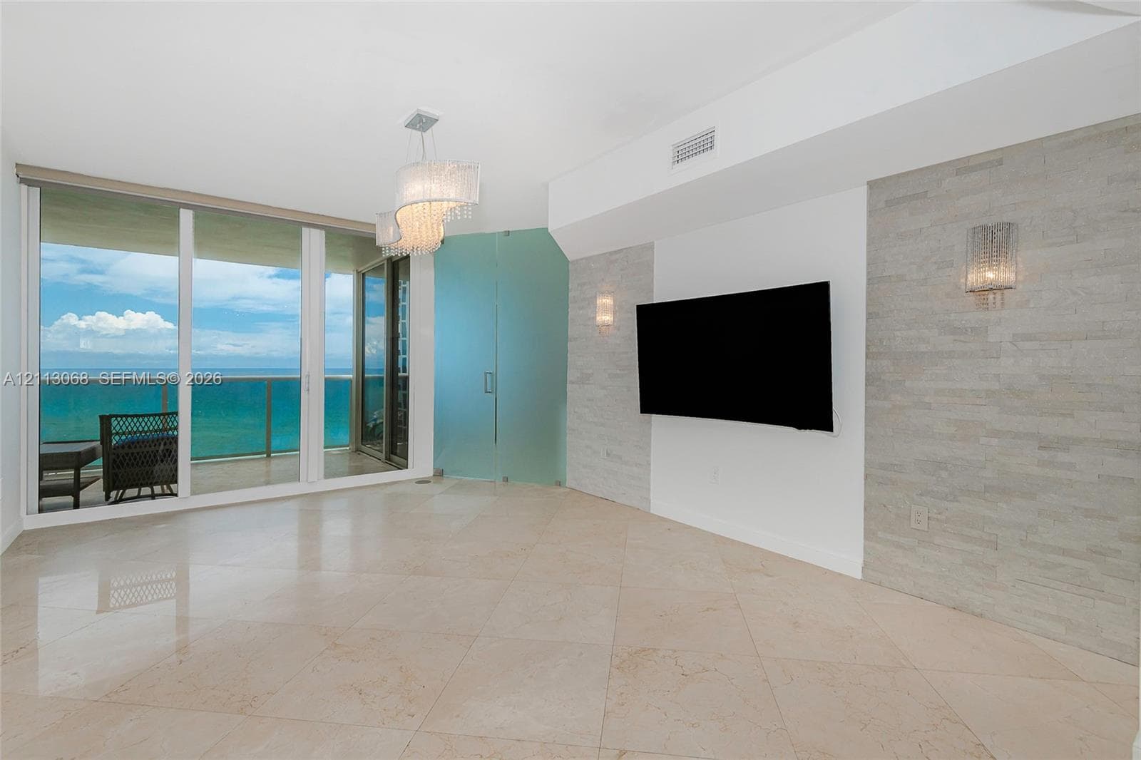 Floor-to-ceiling sliding glass doors, polished stone tile flooring, stacked stone wall feature, frosted glass entry door, and tiered crystal chandelier