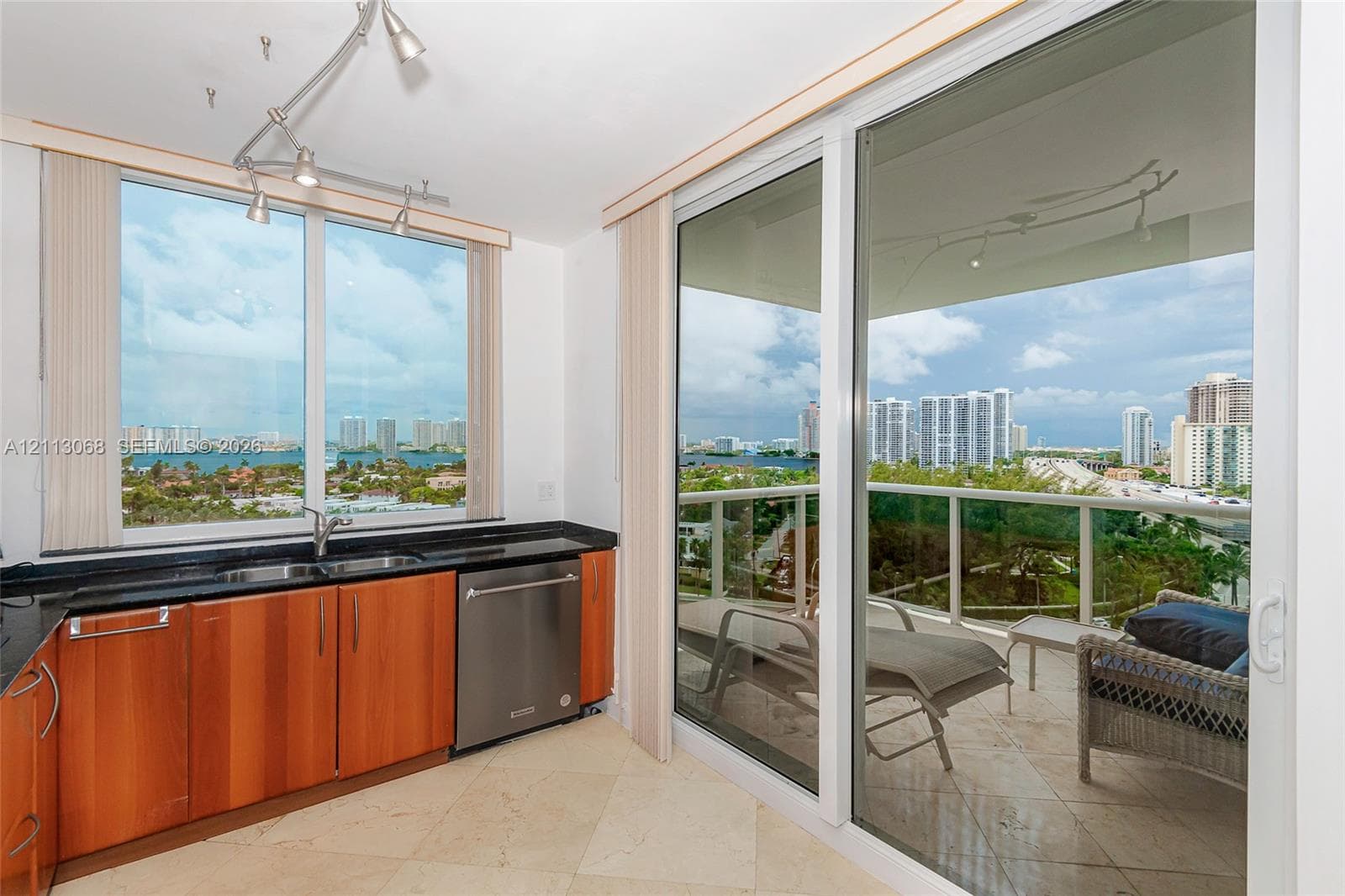 Kitchen area featuring wood-veneer cabinetry, dark stone countertops, stainless steel dishwasher, track lighting, and floor-to-ceiling glass sliding doors opening to a balcony