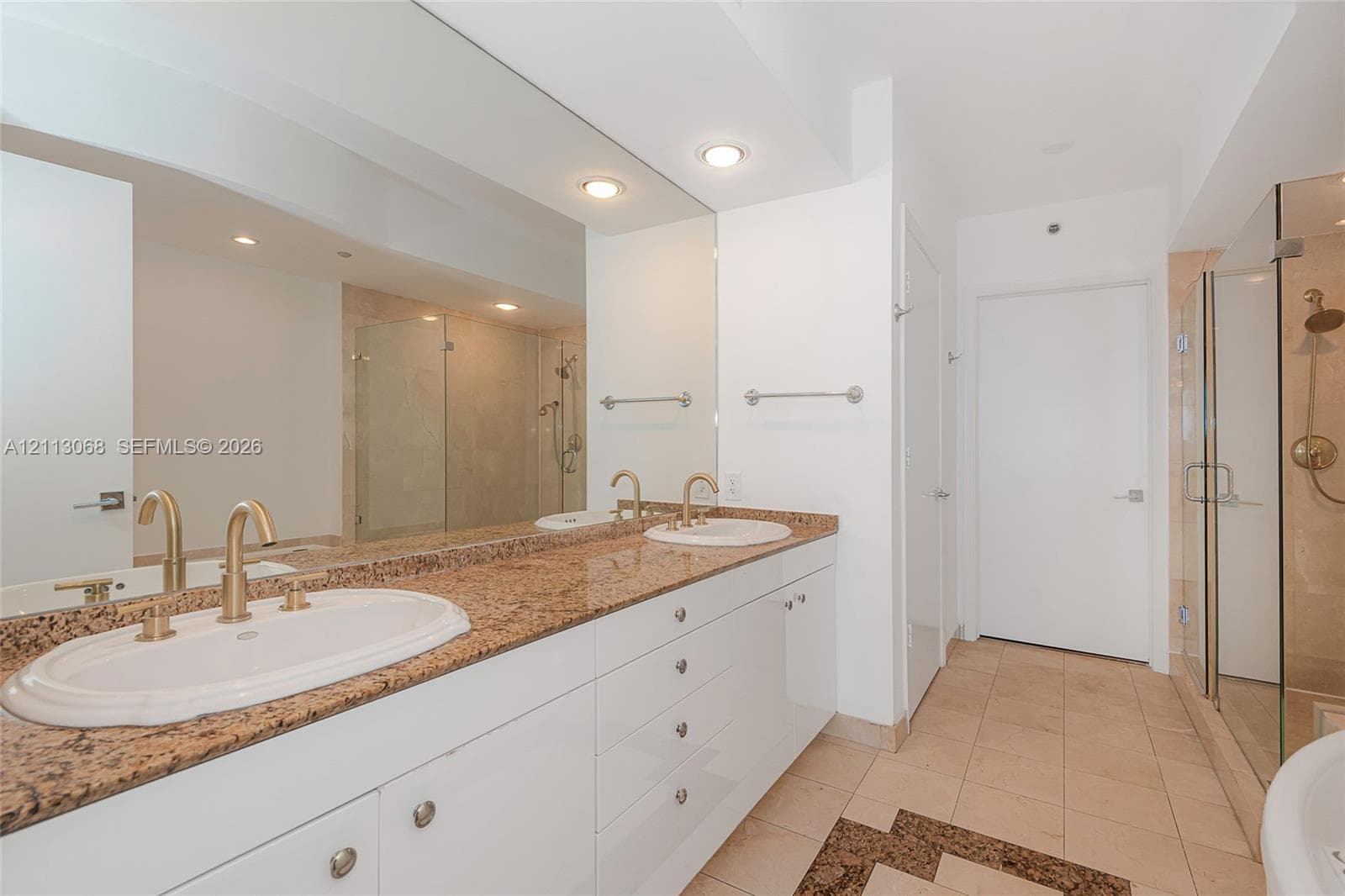 Dual-sink vanity with stone countertop, drop-in basins, brass-toned fixtures, full-width mirror, and a glass-enclosed shower