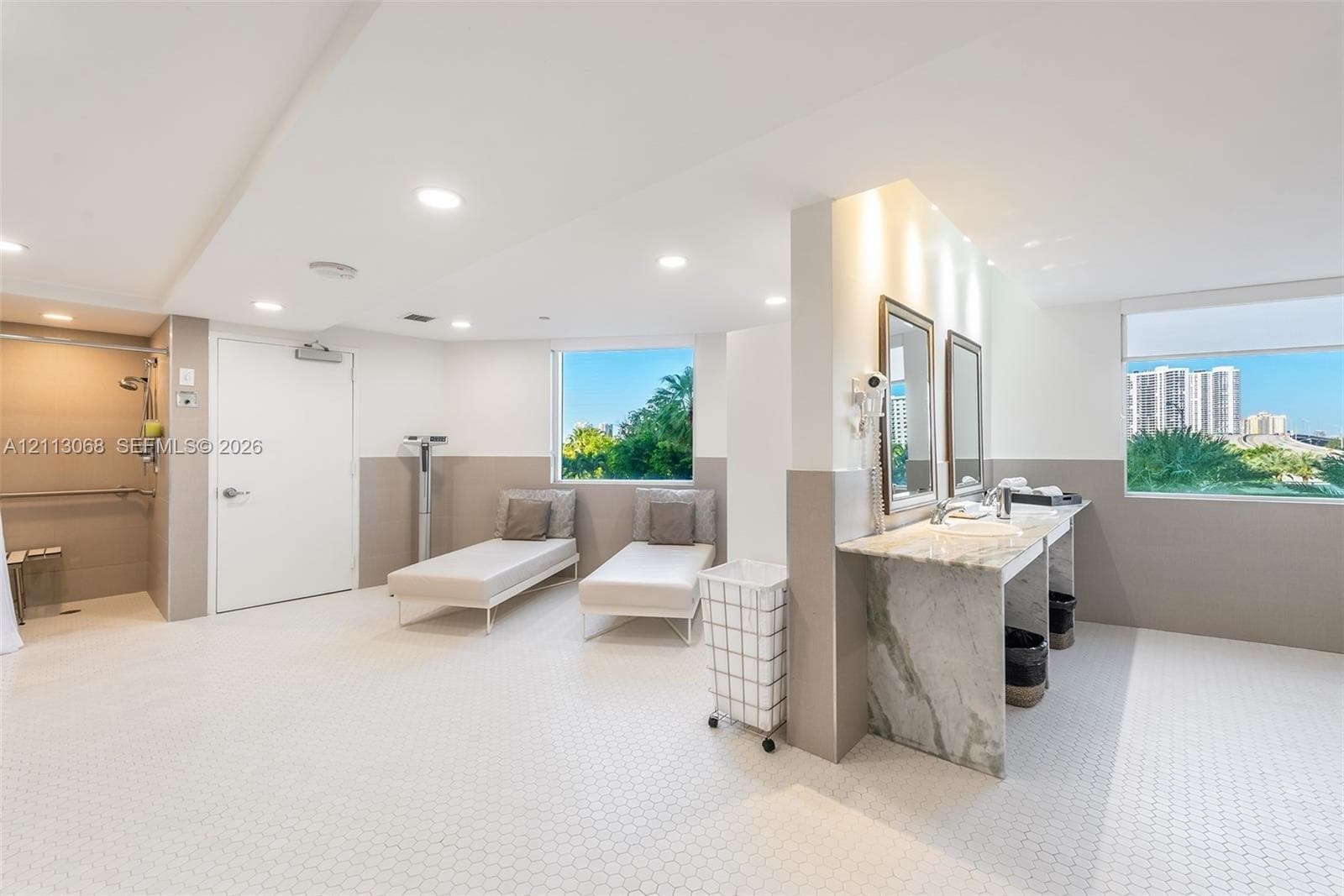 Dual-sink stone countertop vanity, recessed ceiling lighting, tiled flooring, walk-in shower with hand-held fixture, and large windows