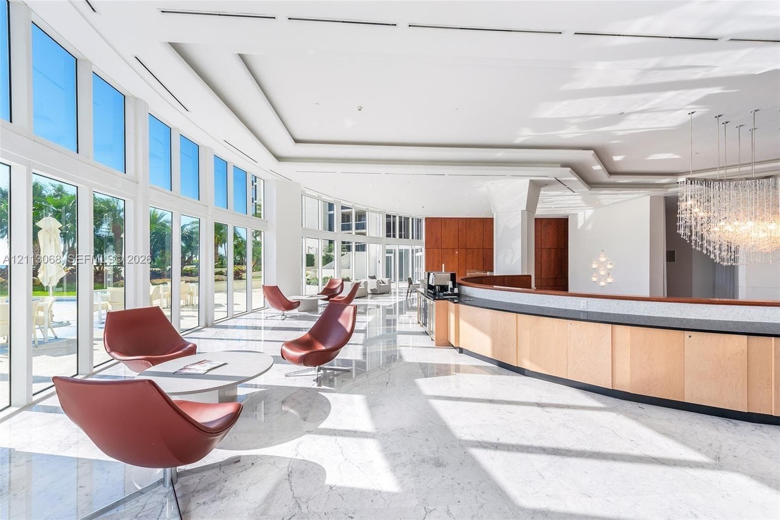 Expansive common space featuring floor-to-ceiling glass panel walls, light-toned stone flooring, tiered tray ceilings, and a large built-in reception desk with a dark stone countertop