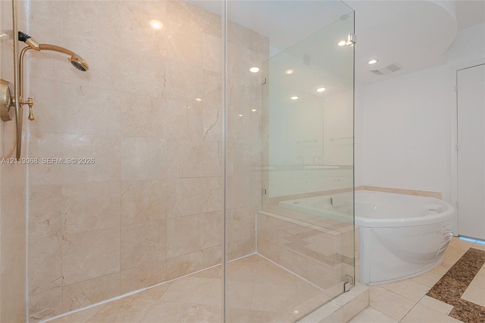 Walk-in shower featuring floor-to-ceiling stone tile, a glass enclosure, and a gold-finish showerhead, paired with a white jetted soaking tub and recessed lighting