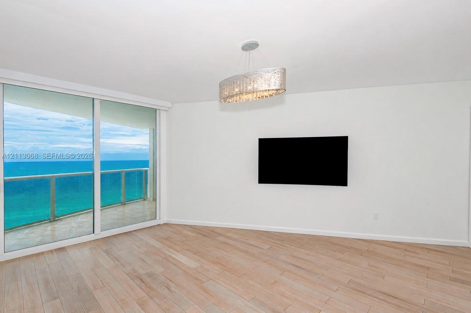 Large sliding glass doors with direct access to a balcony, light-toned wood-finish flooring, oval crystal chandelier, and ocean backdrop