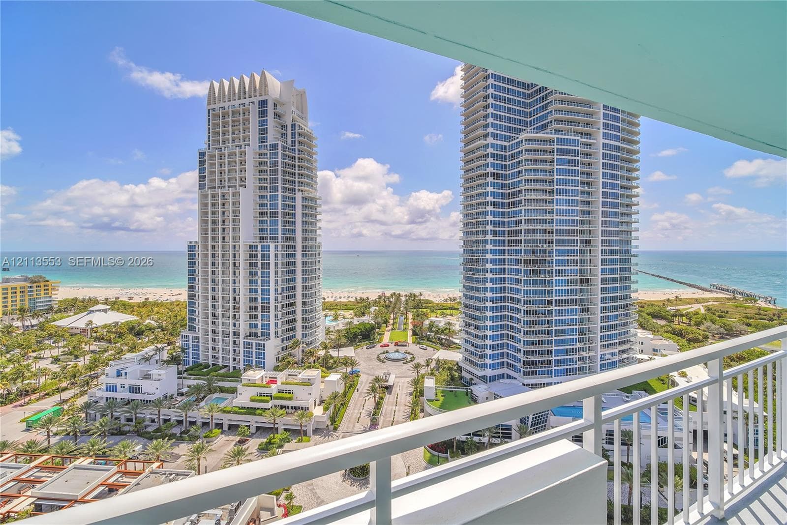 Covered balcony with white metal railings, overlooking coastal high-rise towers, sandy beach, and ocean horizon