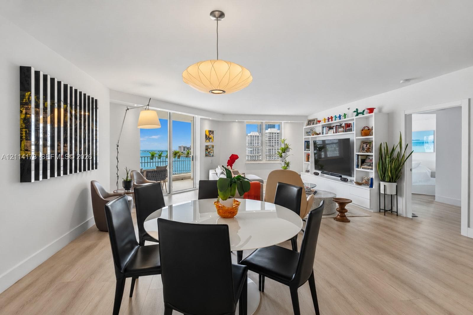 Open-concept interior featuring light wood-finish flooring, floor-to-ceiling glass sliding doors, a custom built-in media wall with integrated shelving, and a prominent saucer-style pendant light