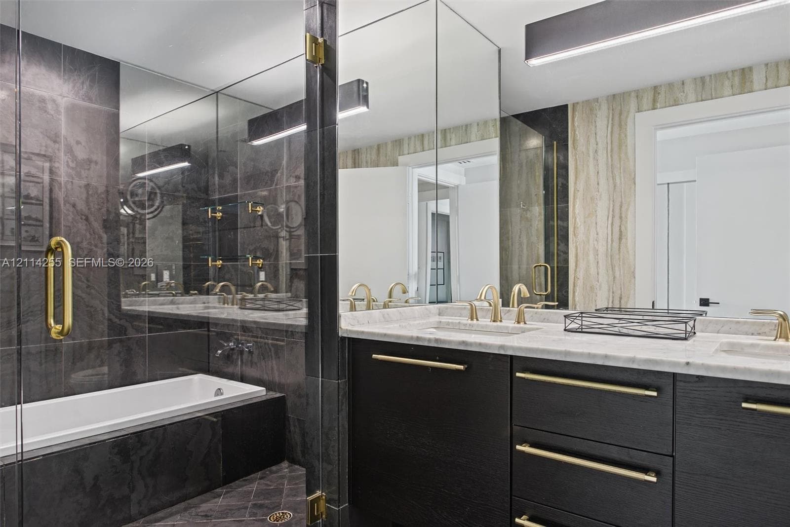 Custom dark wood-finish cabinetry featuring brass hardware, dual undermount sinks, light-toned stone vanity countertop, and a walk-in shower with floor-to-ceiling dark tile surrounds and glass enclosure