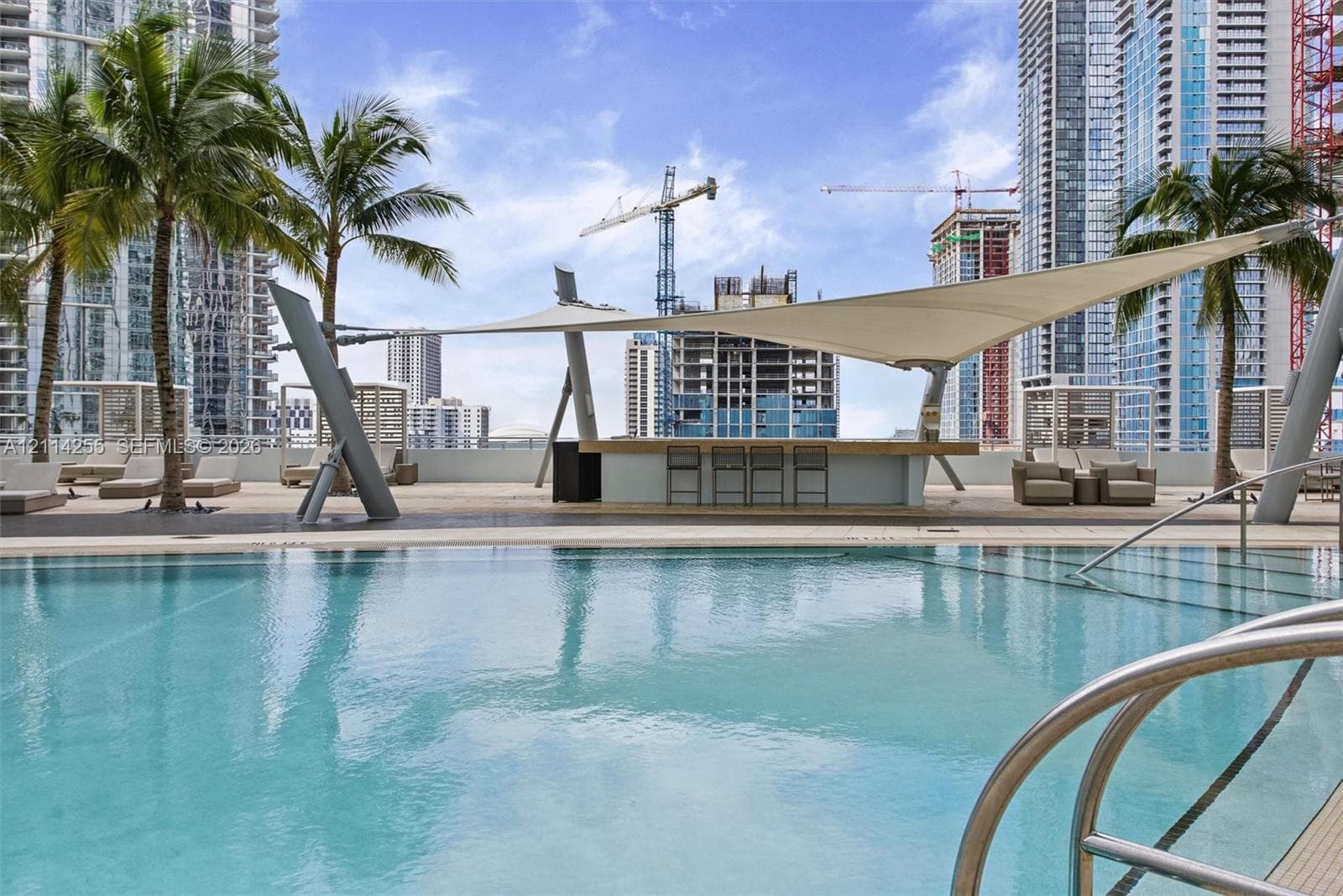 Swimming pool with a stainless steel ladder entry, paved deck, and a built-in bar sheltered by a tensile shade structure