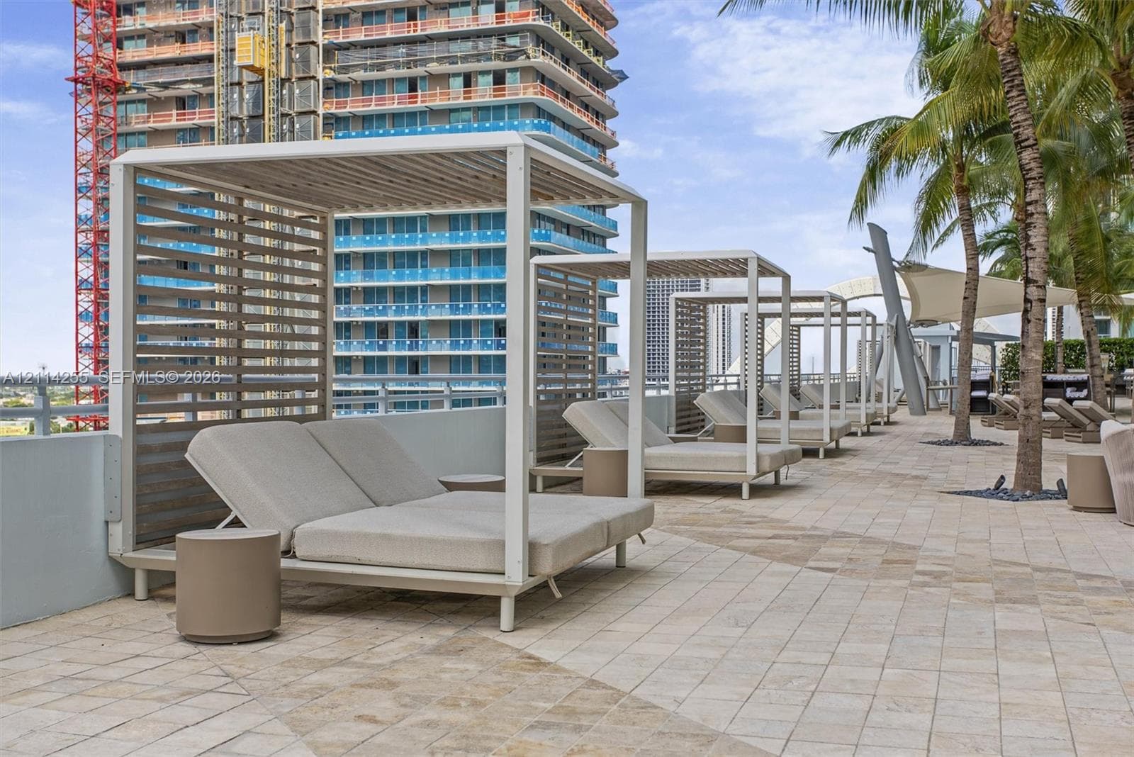 Outdoor terrace featuring light-toned stone tile flooring, structural metal cabanas with slat-wall detailing, and mature palm trees