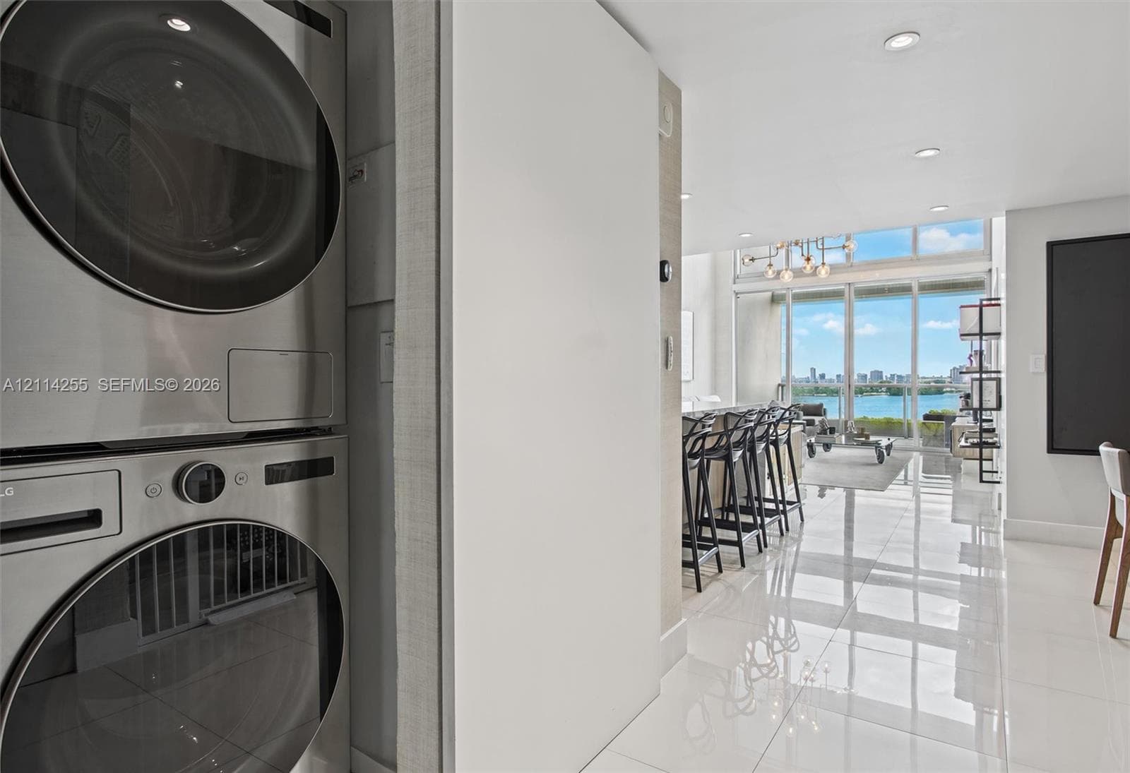 Stacked stainless steel laundry appliances, polished tile flooring, recessed ceiling lighting, and floor-to-ceiling glass paneling